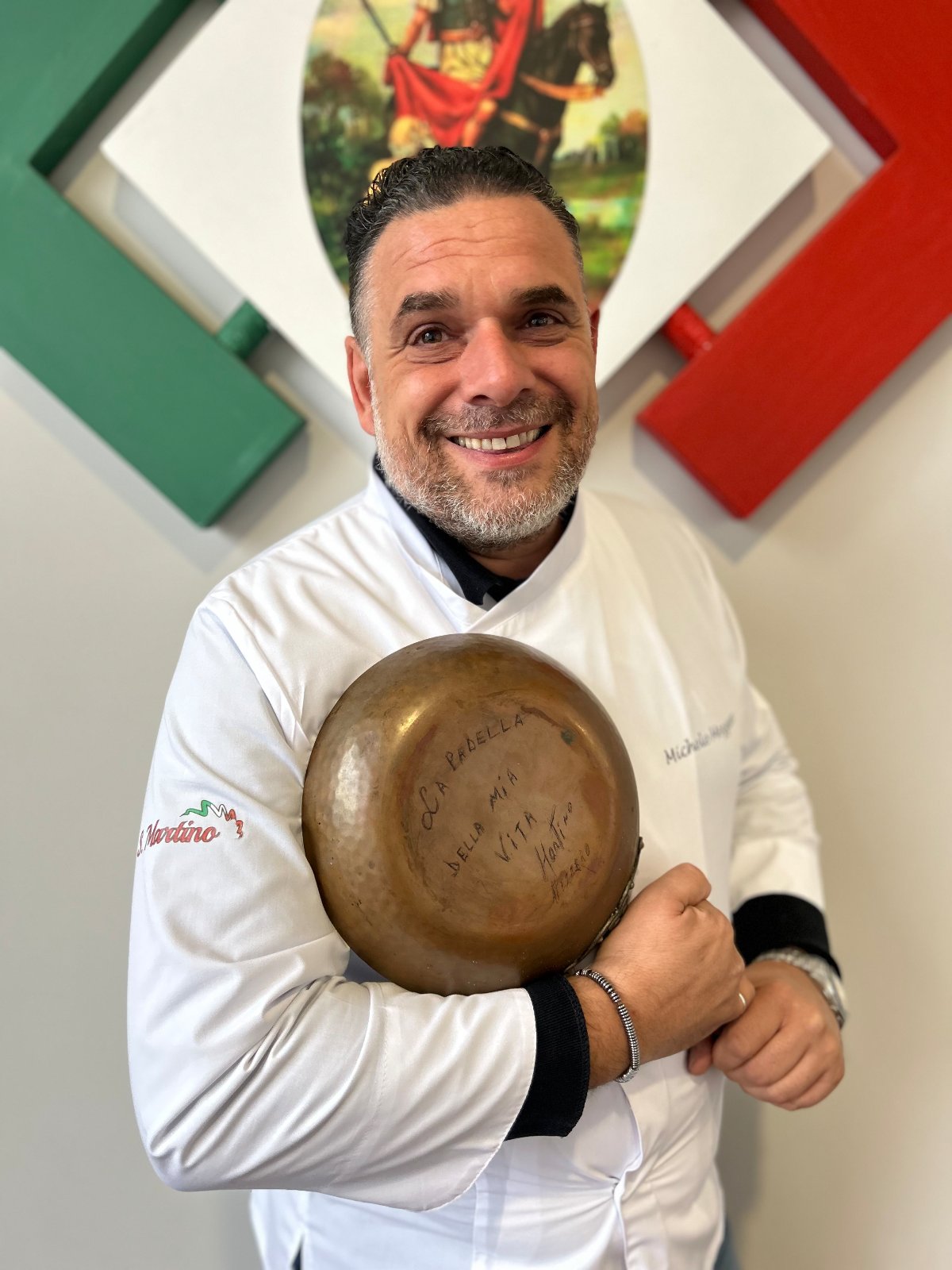 O chef Michele Mezzero sorri com a caçarola de cobre da família nas mãos, diante do quadro de San Martino