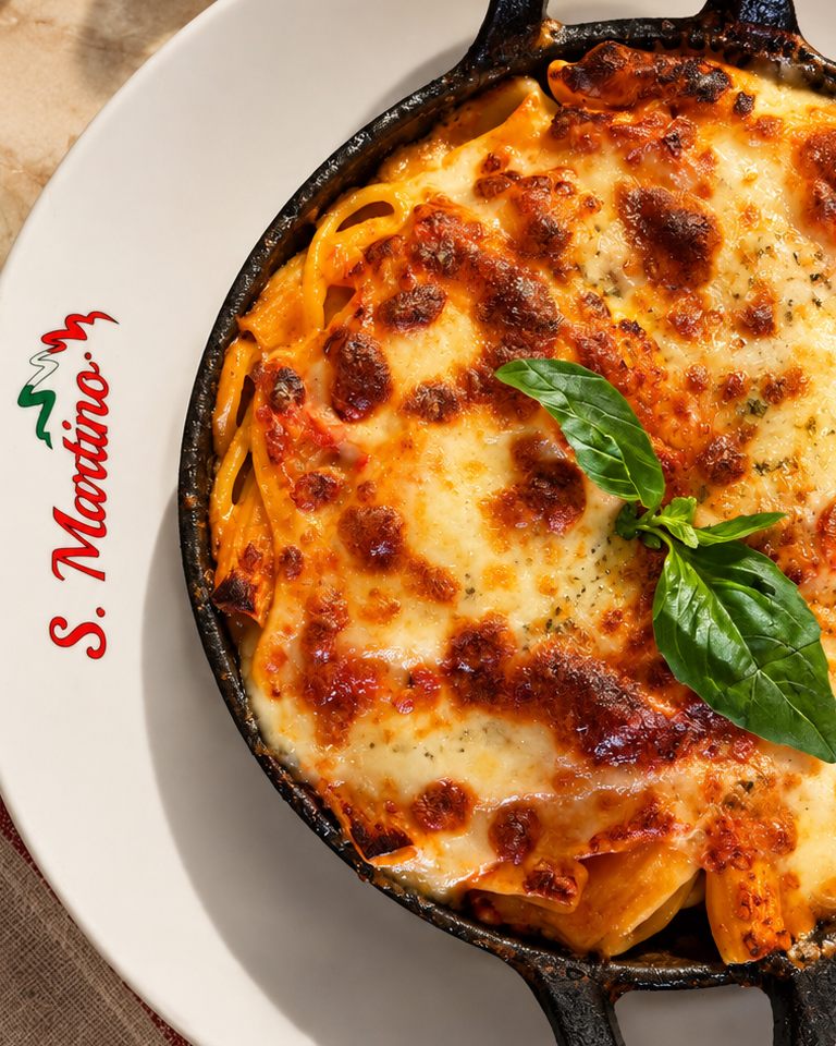 Mistura de massas gratinada com molho bolonhesa, natas e mozzarella numa frigideira de ferro, com manjericão fresco, sobre prato S. Martino