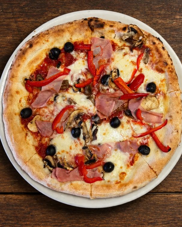 Pizza 4 Stagioni com fiambre, cogumelos, pimento vermelho e azeitonas pretas num prato branco sobre mesa de madeira