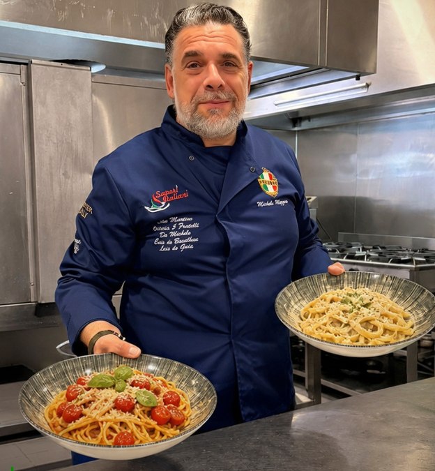 O chef Michele Mezzero na cozinha, de dólman azul, a sorrir e a segurar dois pratos de spaghetti
